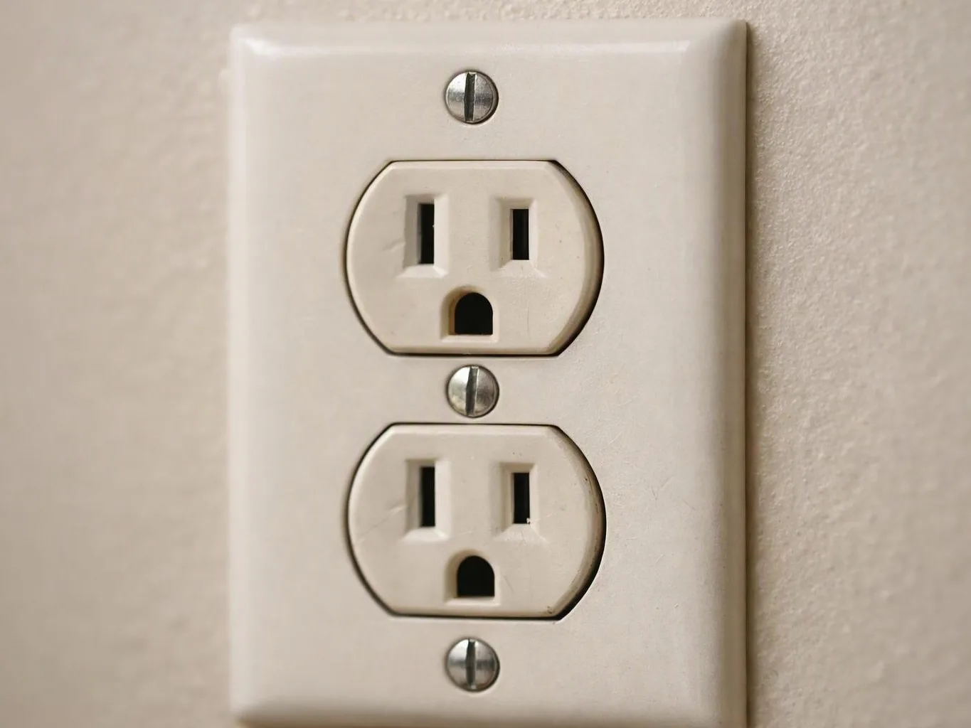 A beige wall outlet with two stacked sockets and visible screw heads sits on a plain indoor wall
