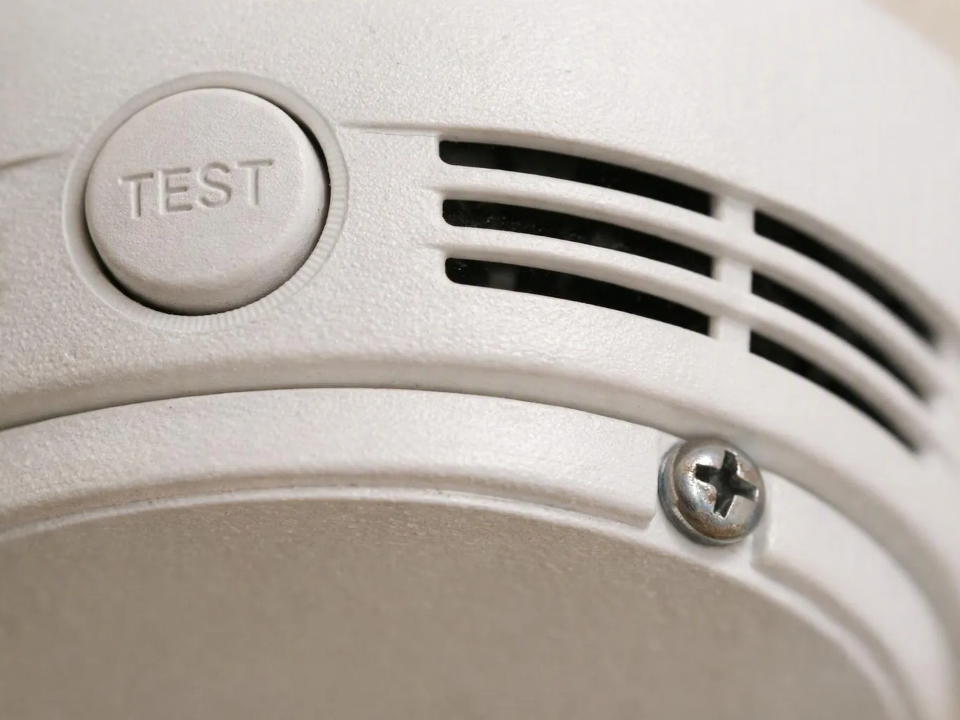 Close-up of a white smoke detector showing a TEST button, vents, and a screw
